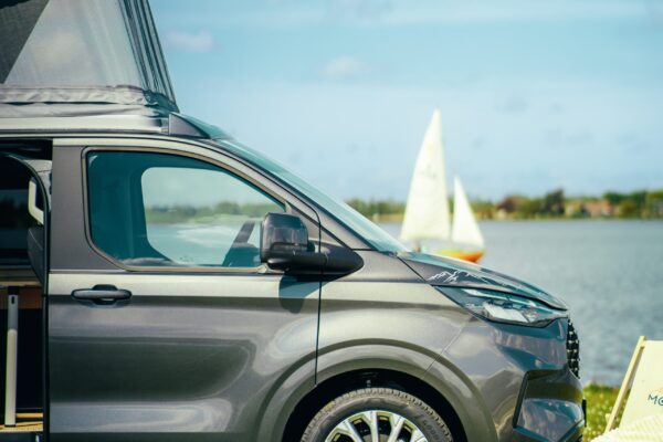 Eine Nahaufnahme des Smarttourer Camper Vans mit aufgeklapptem Aufstelldach. Im Hintergrund ist ein See und auf ihm segelt ein Segelboot. Die silberne Smarttourer-Beklebung glänzt in der Sonne.