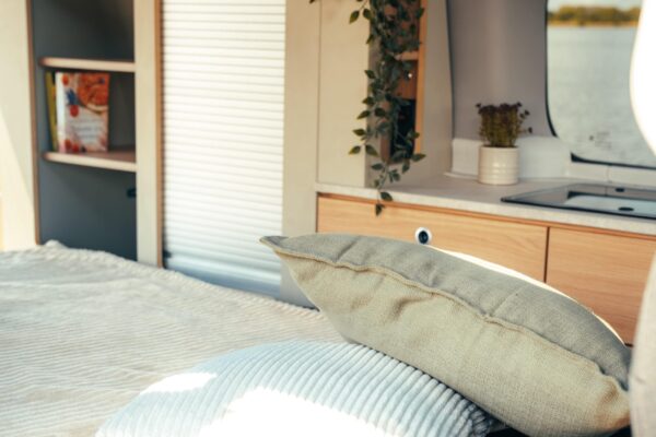 The interior of the Smarttourer camper van. The folded-out Smartbench transforms the inside of the camper van into a cozy bedroom in no time at all. There is a blanket and two soft pillows on the sleeping area. The open cupboard module with books and other items can be pulled out in the background. You can see the water through the window.