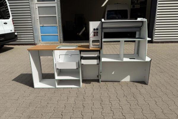 Das Smartkit Leichtbaumöbelset für den Selbstausbau des eigenen Camper Vans. Graue Fronten mit Schubladen und Schränken. Eine Arbeitsplatte in Holzoptik. Außerdem gibt es Aussparungen für den Kühlschrank und das Spülbecken. Abgebildet wird das Set von hinten. Man erkennt die Stromanschlüsse.